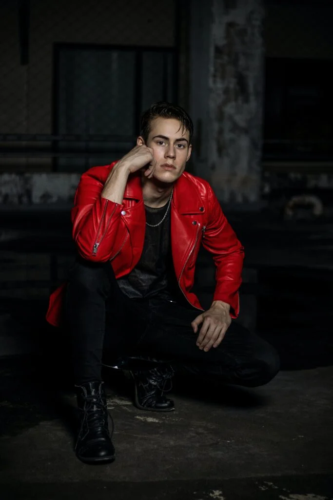 A fashion model wearing a red leather jacket, a chain, and black leather boots. He is donning a bold and edgy look with the vibrant red leather jacket and the stylish chain. The black leather boots complete the ensemble, adding a touch of elegance and sophistication. Click to shop our Men's page and discover more fashion-forward clothing and accessories to elevate your wardrobe.