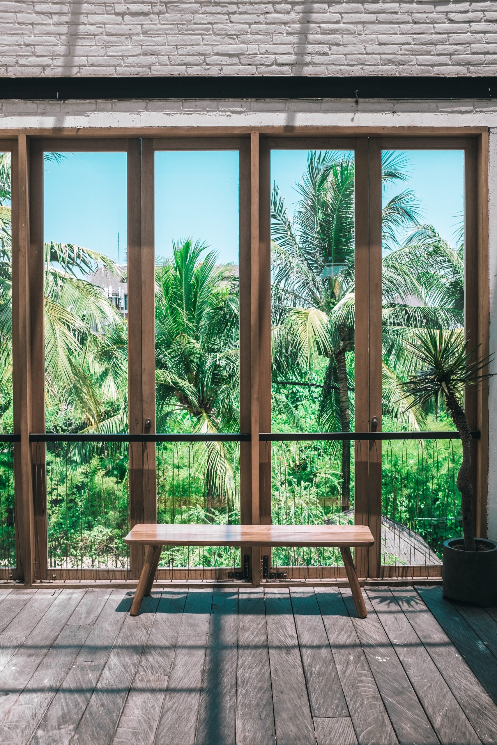 A floor to ceiling glass window frame with palm vegetation in the background and a resting bench on the wooded floor