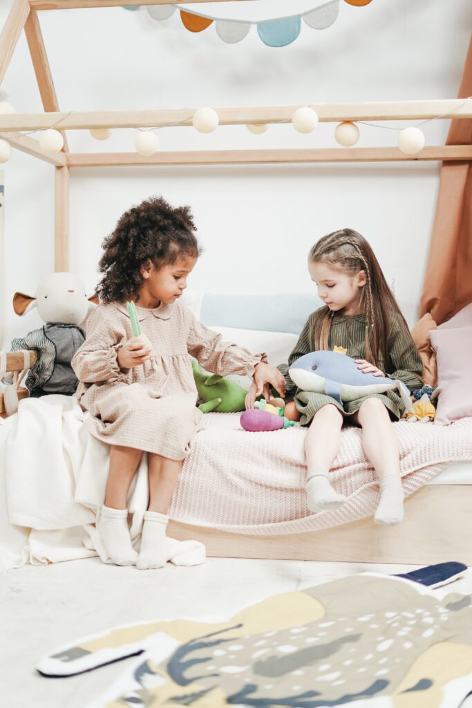 Two girls playing with dolls on a bed in a serene environment and wearing high quality clothing pieces